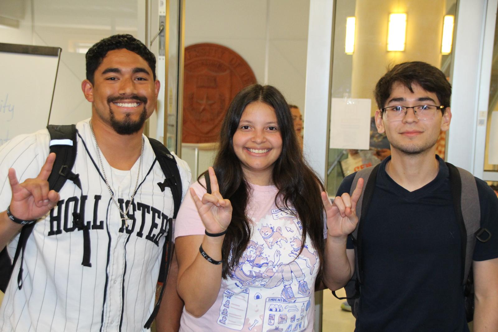 Three first-gen students smiling happily holding up a hook 'em horns symbol at an event. 