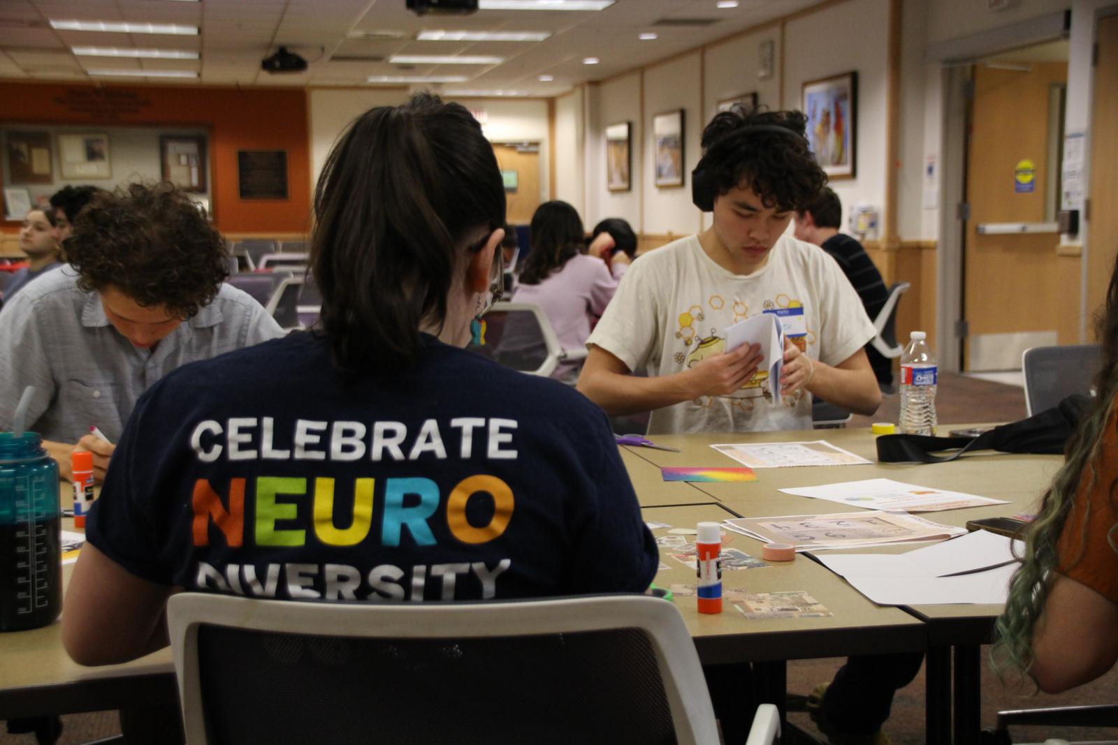 Students sitting at a desk and crafting pieces of paper. In the foreground, there is a staff member with a "celebrate neurodiversity" shirt written on the back.