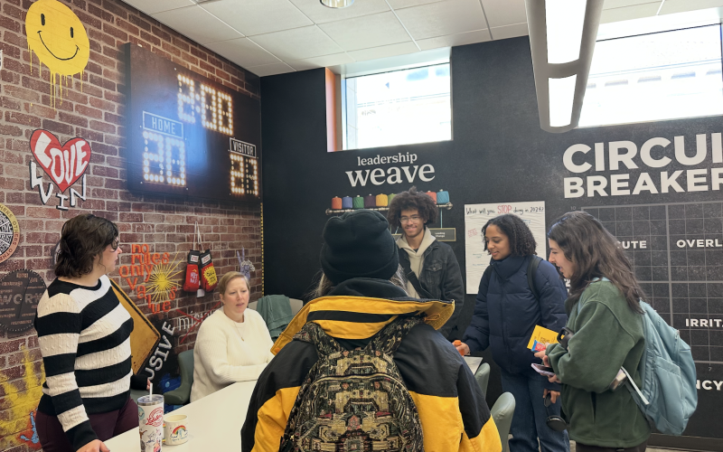 students gathered around a table looking at staff who are speaking to them. The room is filled with objects such as a fake basketball scoreboard and a smiley face poster on a brick wall