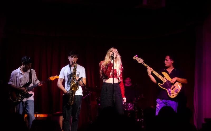 band standing on a stage and performing. Lead singer is singing, while two musicians play the guitar and one plays the saxophone